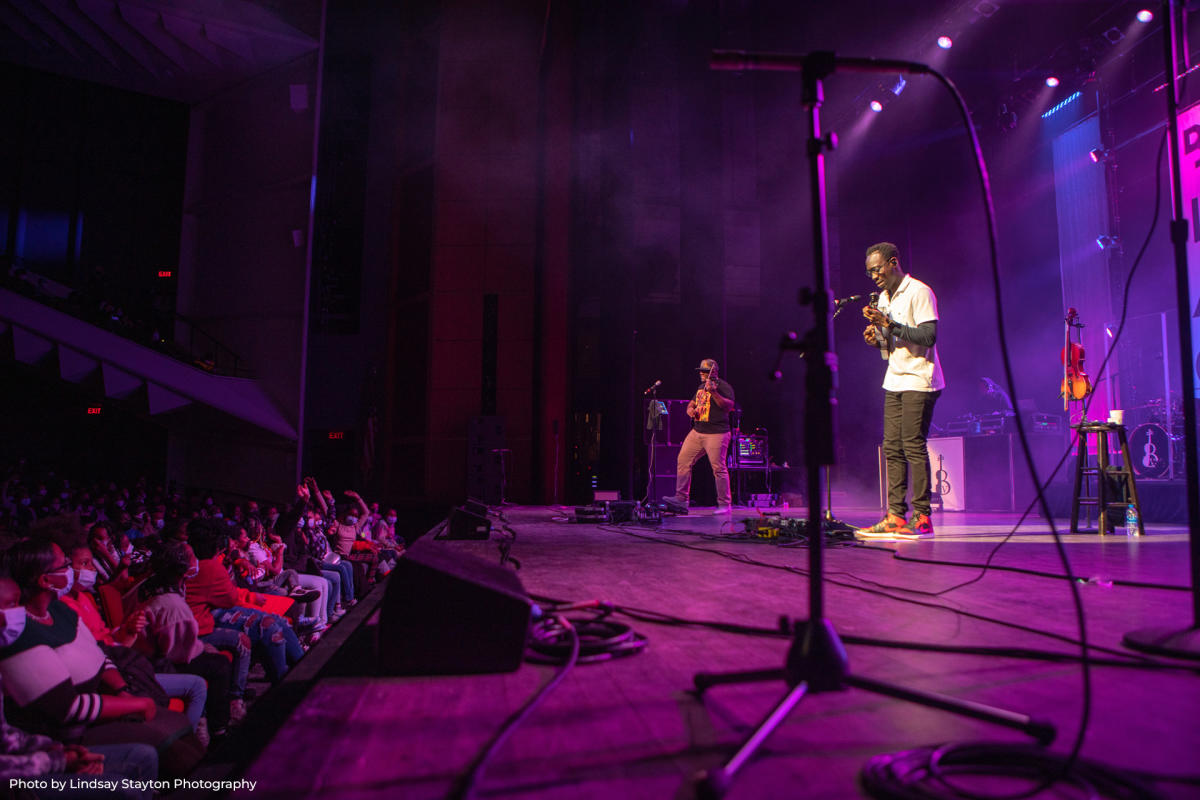 A live concert performance on stage with two musicians playing string instruments under purple lighting, while a large audience watches from theater seating. One musician plays at the front of the stage, another stands further back, and a violin rests on a stool nearby.