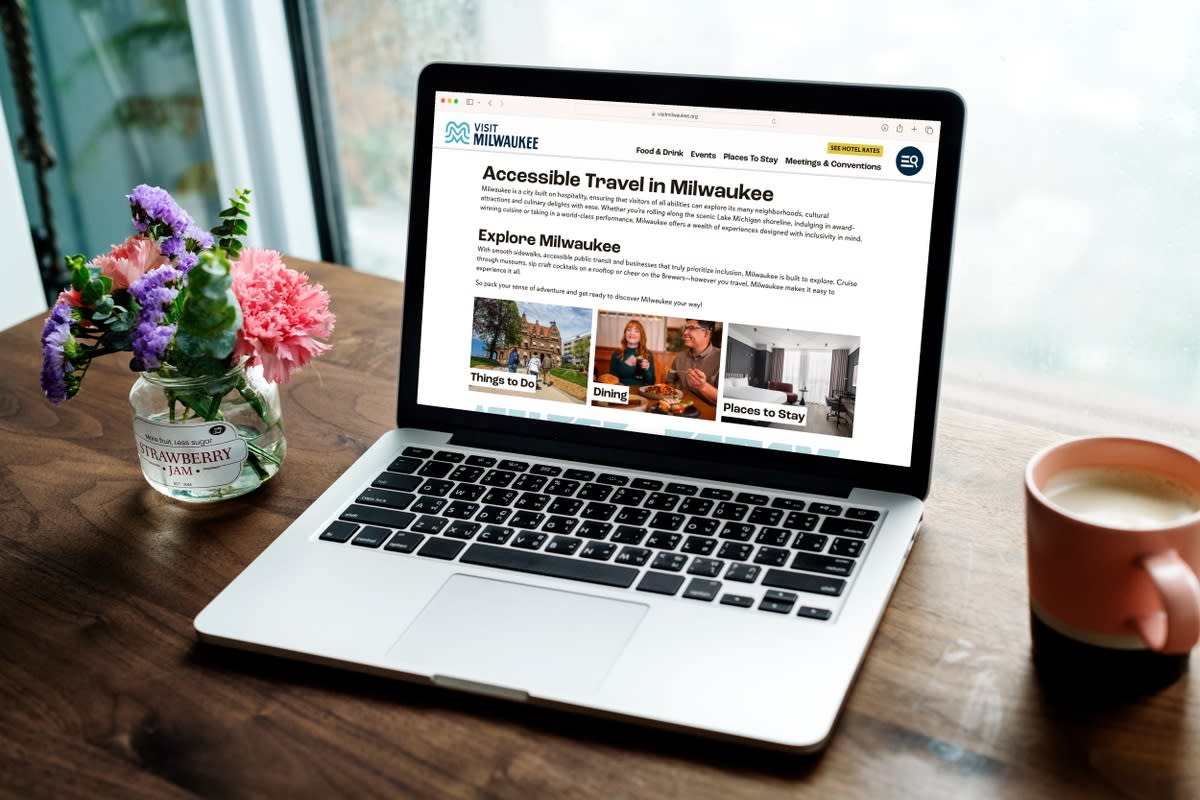 A laptop on a wooden table displaying the "Accessible Travel in Milwaukee" webpage from Visit Milwaukee. The screen features sections titled "Things to Do," "Dining," and "Places to Stay." Next to the laptop is a small bouquet of flowers in a glass jar labeled "STRAWBERRY JAM" and a mug of coffee. A large window with raindrops in the background suggests a cozy indoor setting.