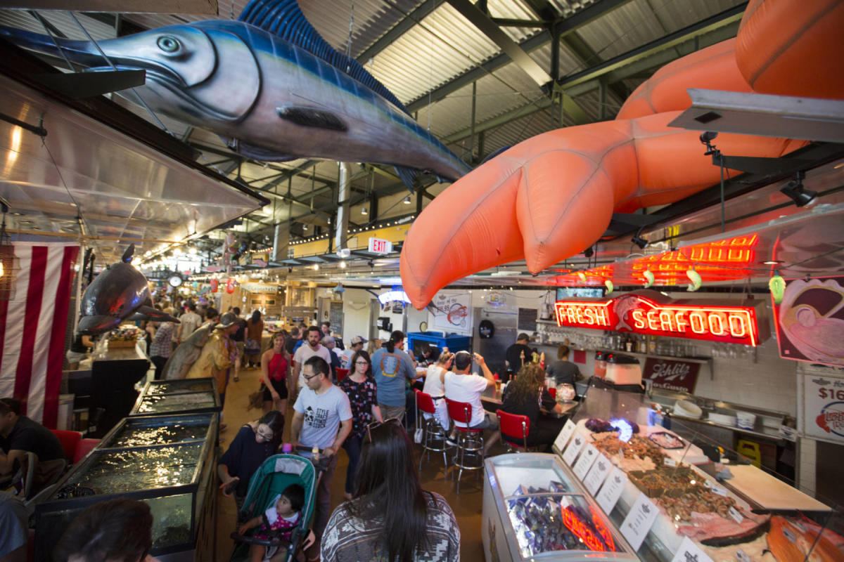A bustling indoor seafood market filled with people browsing and dining, featuring large hanging fish decorations, bright neon “Fresh Seafood” signs, and displays of fresh catches on ice.
