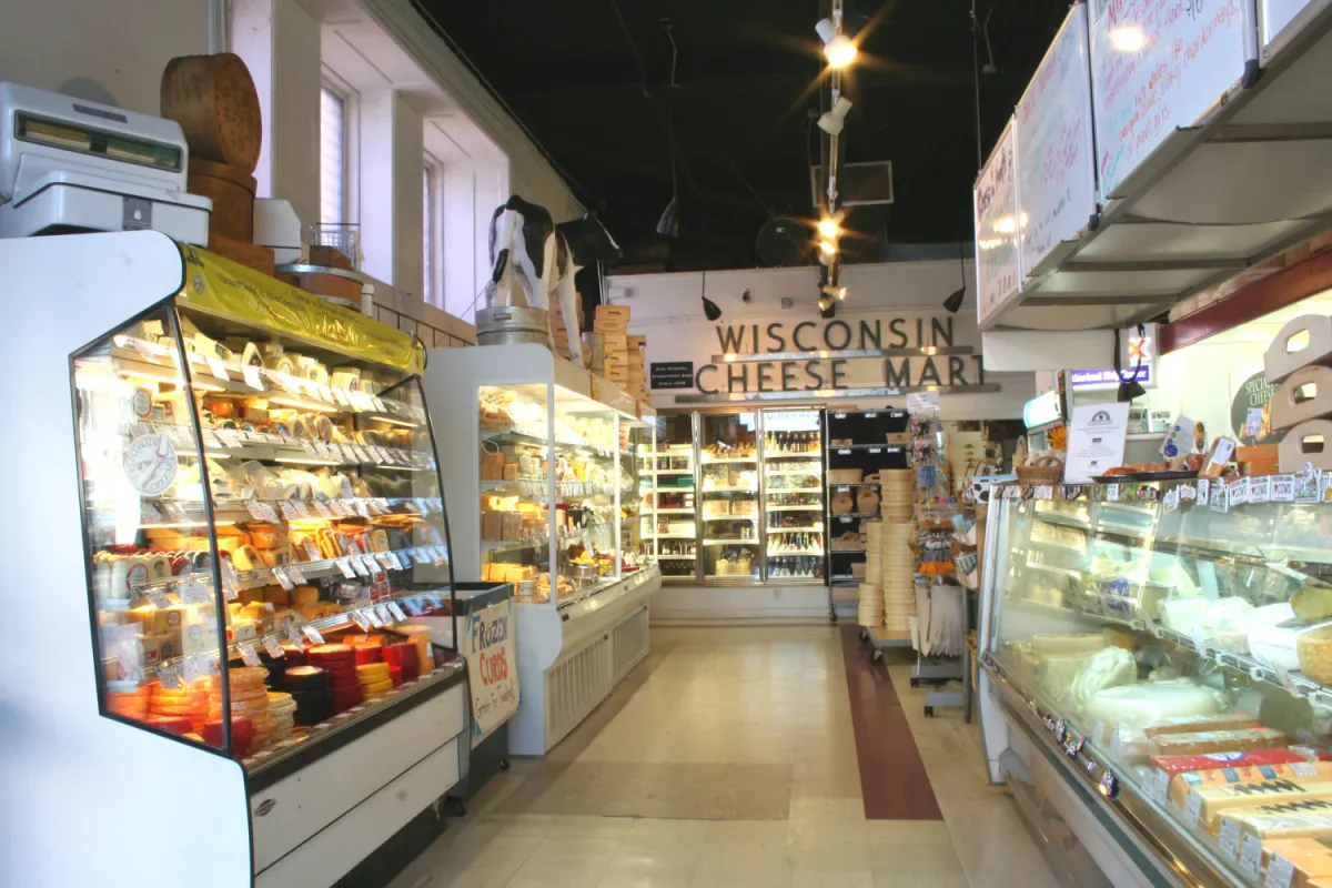 Interior of a cheese shop with refrigerated display cases filled with cheese and dairy products, bright lighting, and a sign in the back reading “Wisconsin Cheese Mart.”