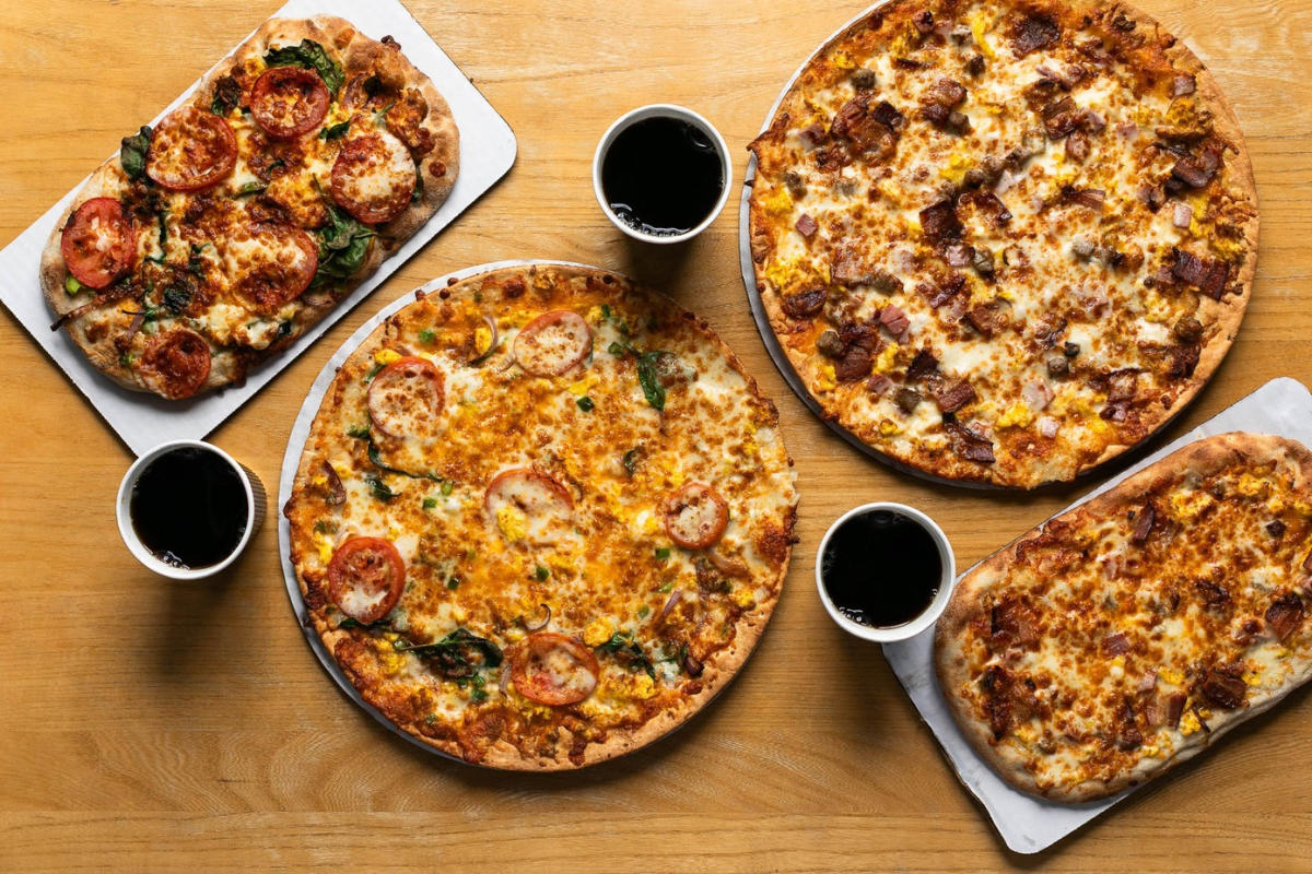 Overhead view of four freshly baked pizzas on a wooden table, including round and oval pies topped with melted cheese, tomatoes, herbs and bacon, with cups of dark soda placed between them.