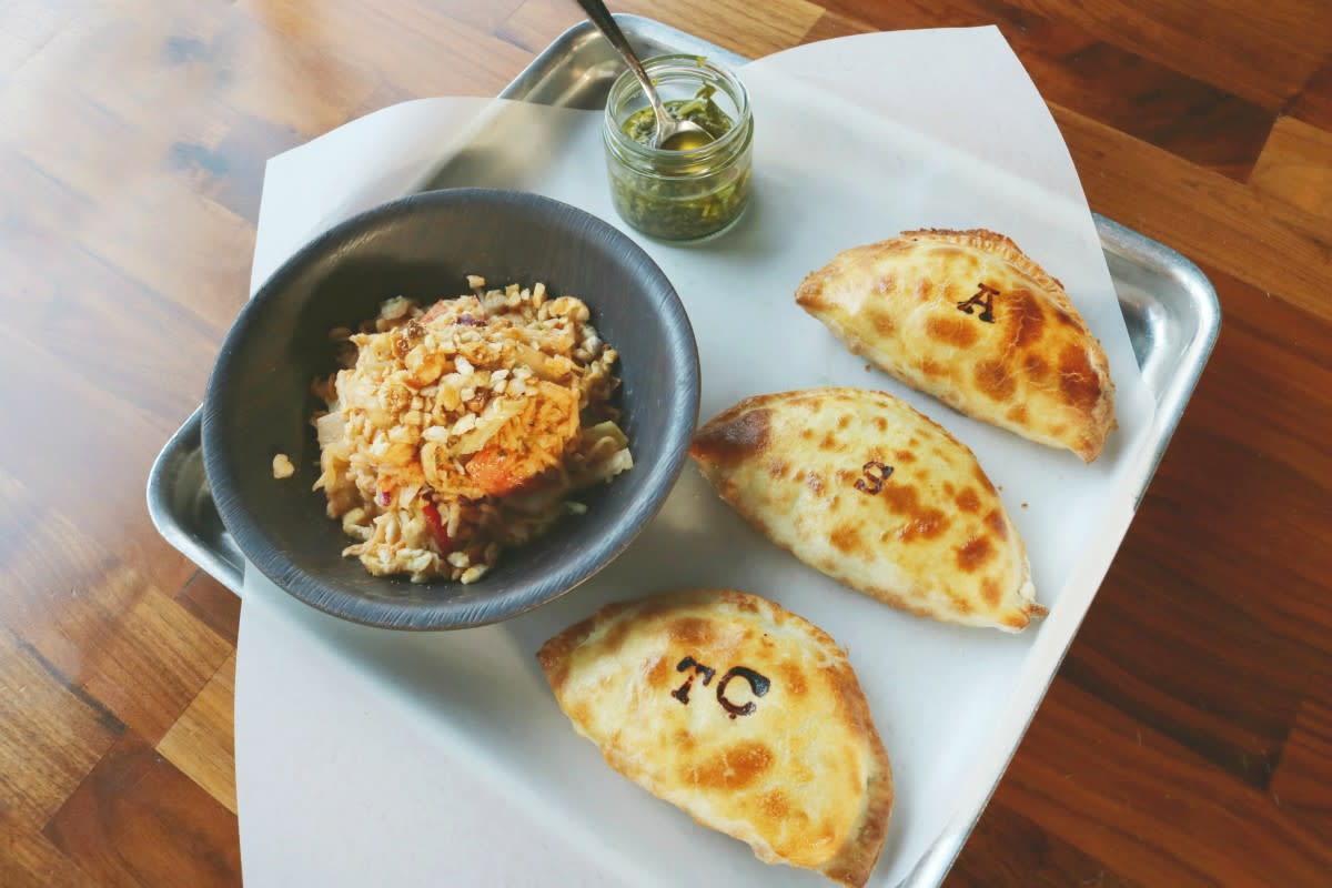 A tray with three golden-brown empanadas, each stamped with different initials, served alongside a bowl of seasoned rice topped with crispy bits and a small jar of green sauce.