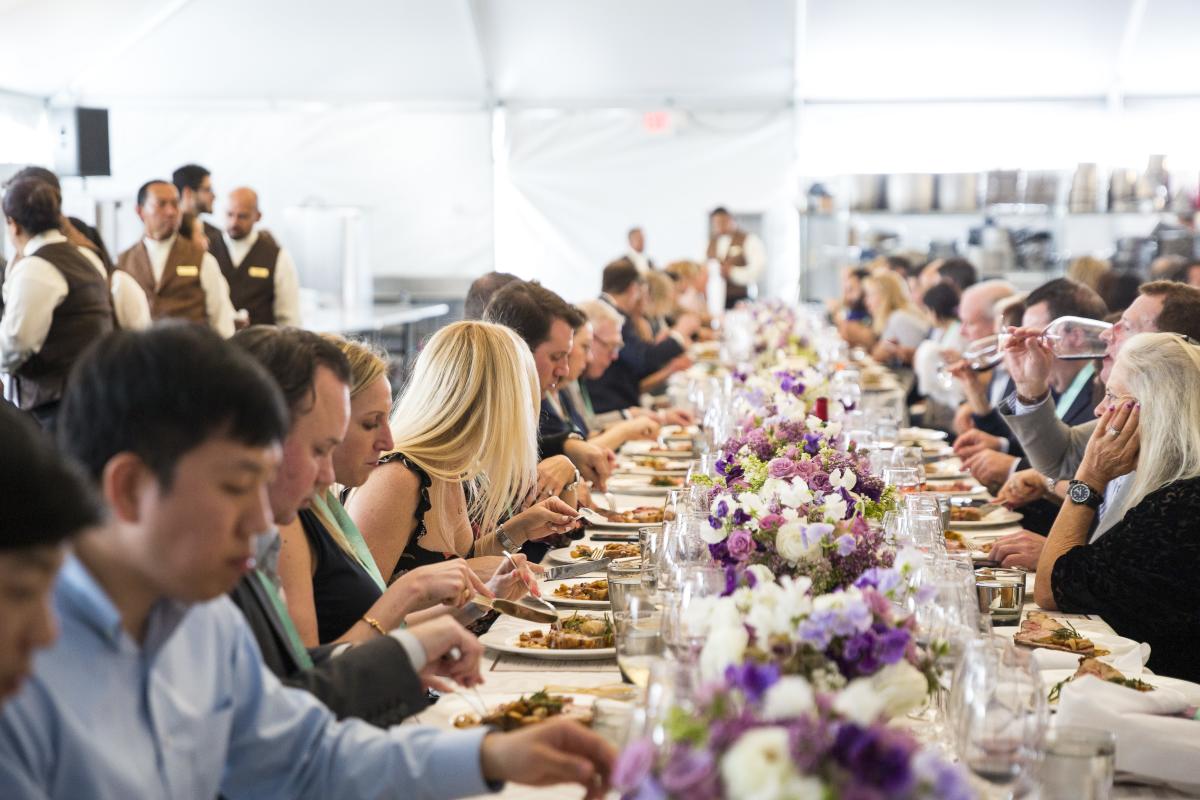 This is an image of a decorated table of guests enjoying a meal and wine at Pebble Beach Food & Wine. The table is lined with colorful bouquets of purple, white and green florals