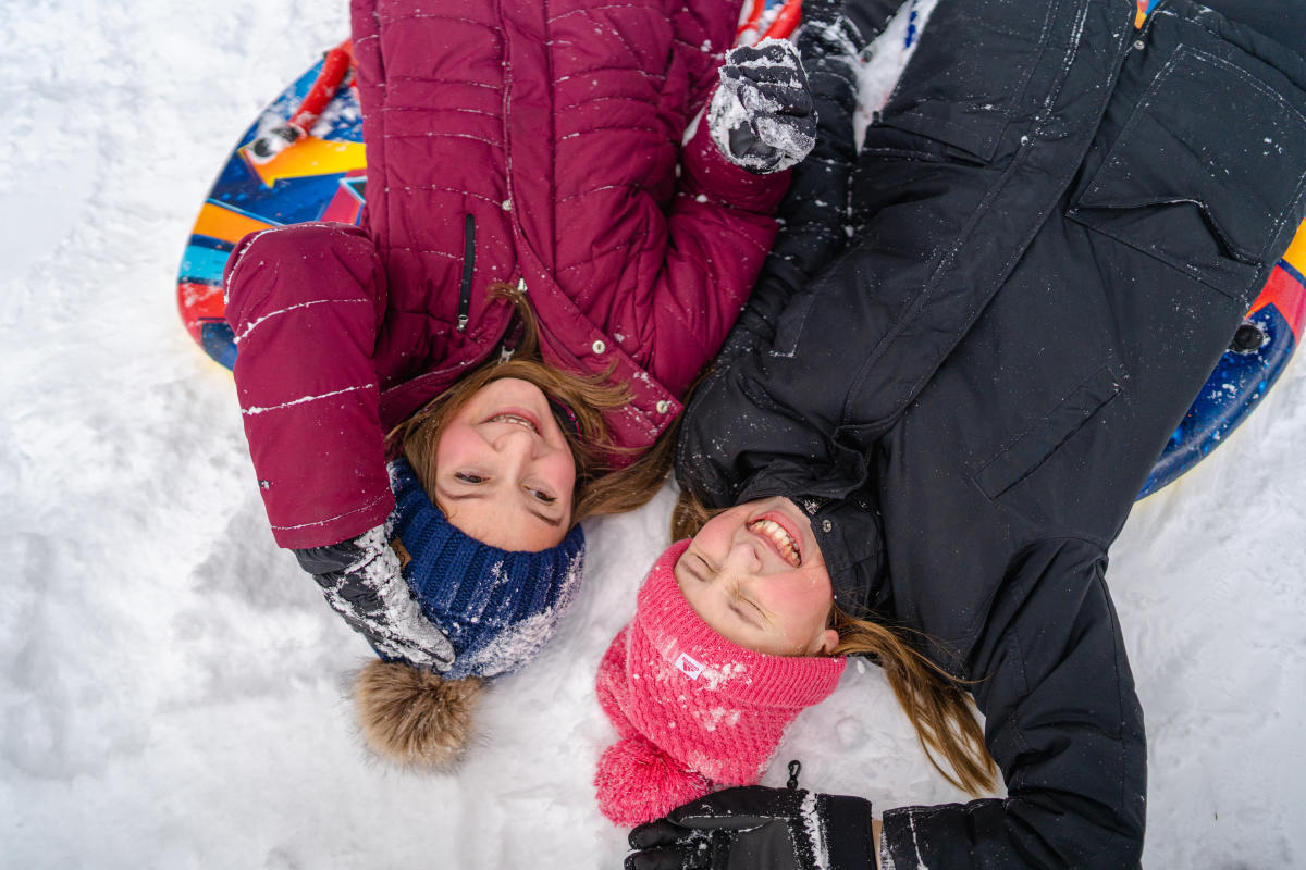 2 young girls in winter wear lay laughing in snow with sleds