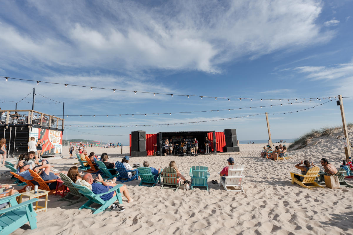 Outdoor concert on a sandy beach at The Deck with people sitting on colorful chairs under a clear sky, enjoying live music from a band performing on a black and red stage.