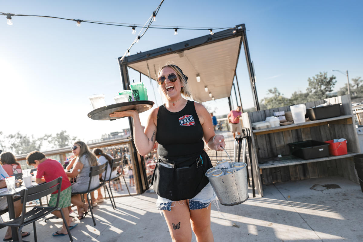 Smiling waitress holds bucket of iced drinks and tray of drinks in outdoor beach setting restaurant.