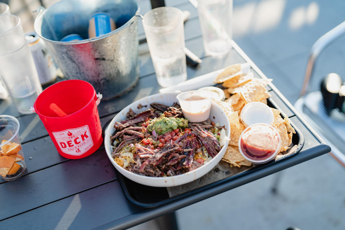 A meal of shredded bbq, guacamole, and sauces served on a tray with a drink bucket labeled "The Deck" and empty glasses on a table.