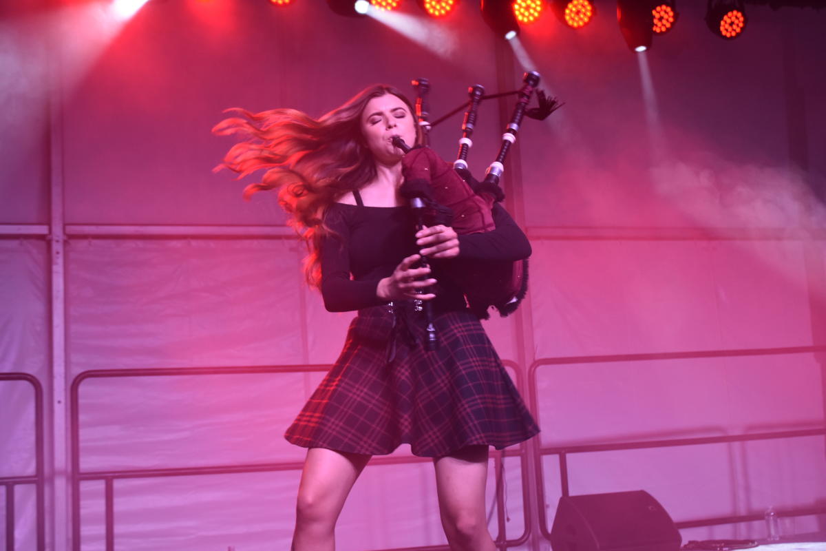 Performer playing bagpipes on stage at an event, wearing a tartan skirt and moving energetically under purple stage lighting.