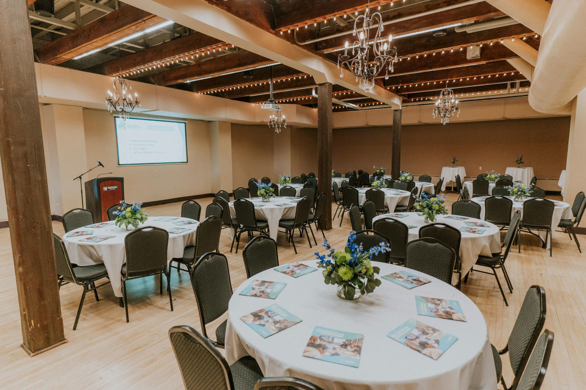 A well-lit banquet hall with round tables set in rows, adorned with white tablecloths, blue floral centerpieces, and event programs. Elegant chandeliers hang from the ceiling.