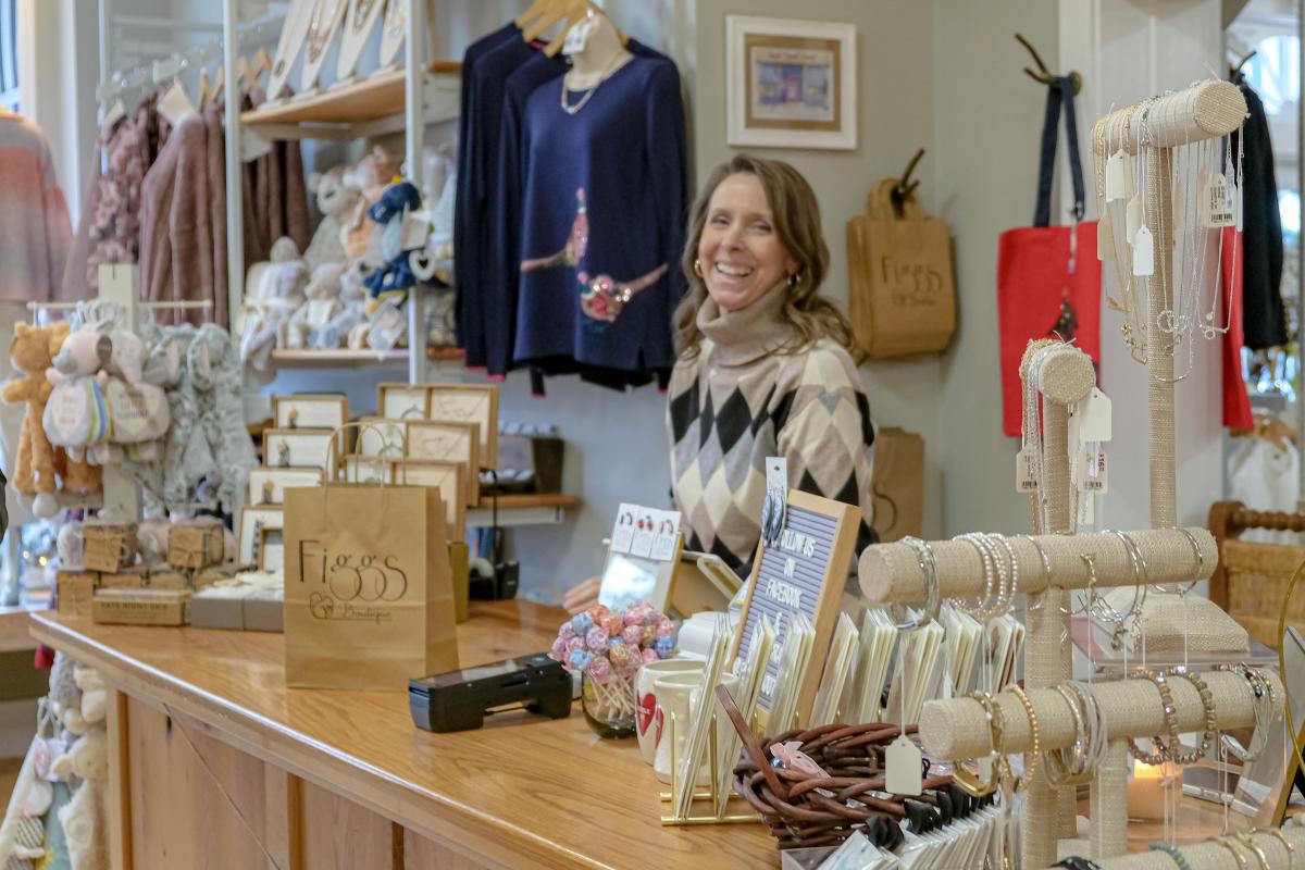 A person smiling behind the cashier counter in a boutique store called "Figgs Boutique." The store is well-stocked with various clothing items, accessories, and unique gifts displayed neatly around the shop.