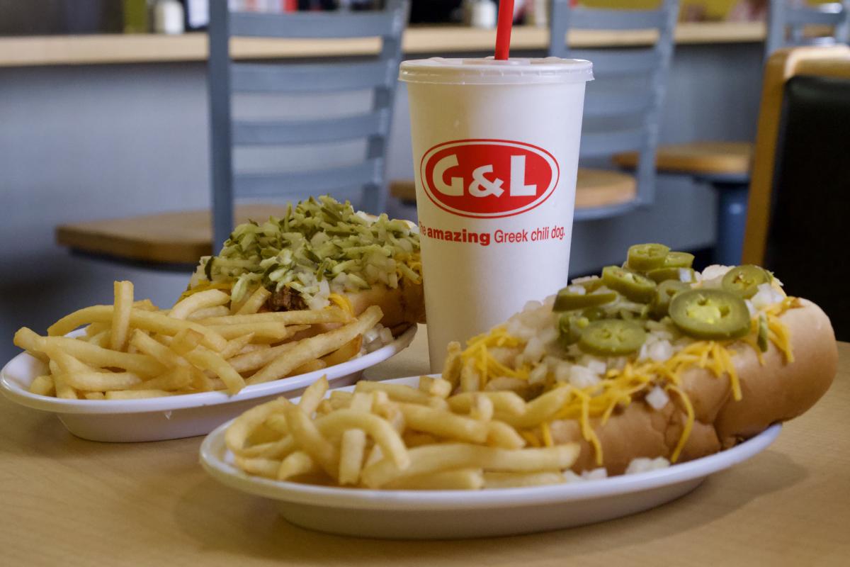 A meal from G&L featuring a chili dog topped with cheese and jalapeños, a burger with pickles, and two portions of French fries, alongside a branded G&L drink cup with a straw, set on a wooden table.