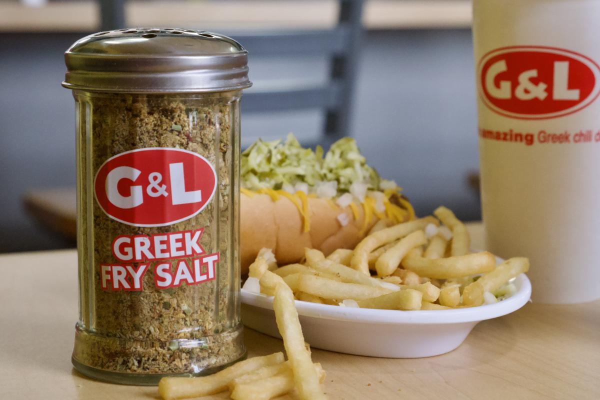 A product labeled "G&L Greek Fry Salt" alongside a hot dog topped with relish and a side of fries, with a cup marked "G&L Amazing Greek Chili Dogs" in the background.