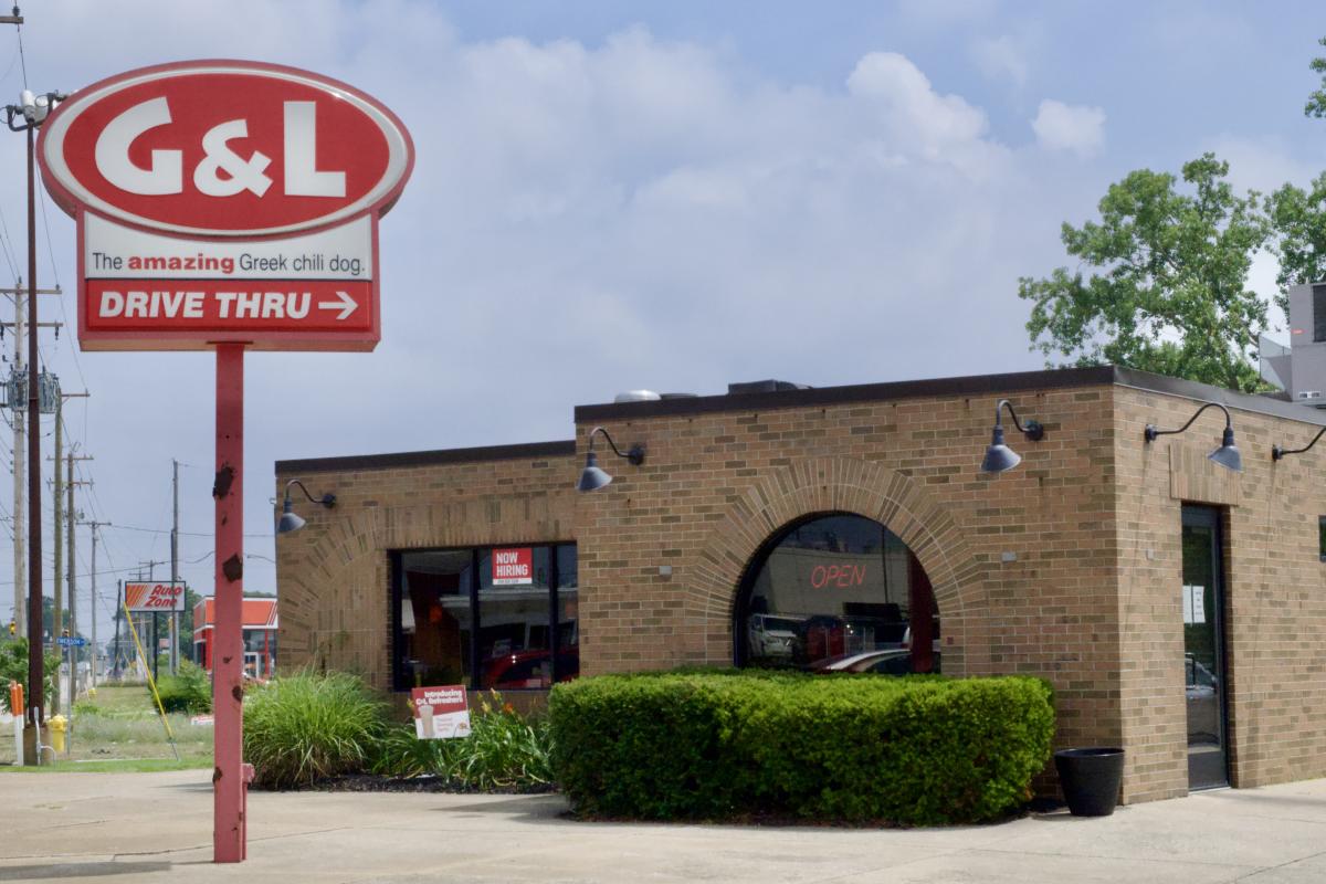 Exterior view of G&L Chili Dogs restaurant with a prominent red sign featuring the logo and a directional arrow for a drive-thru. The building is brick with large windows and an "OPEN" sign displayed. There are landscaping bushes in front of the building.