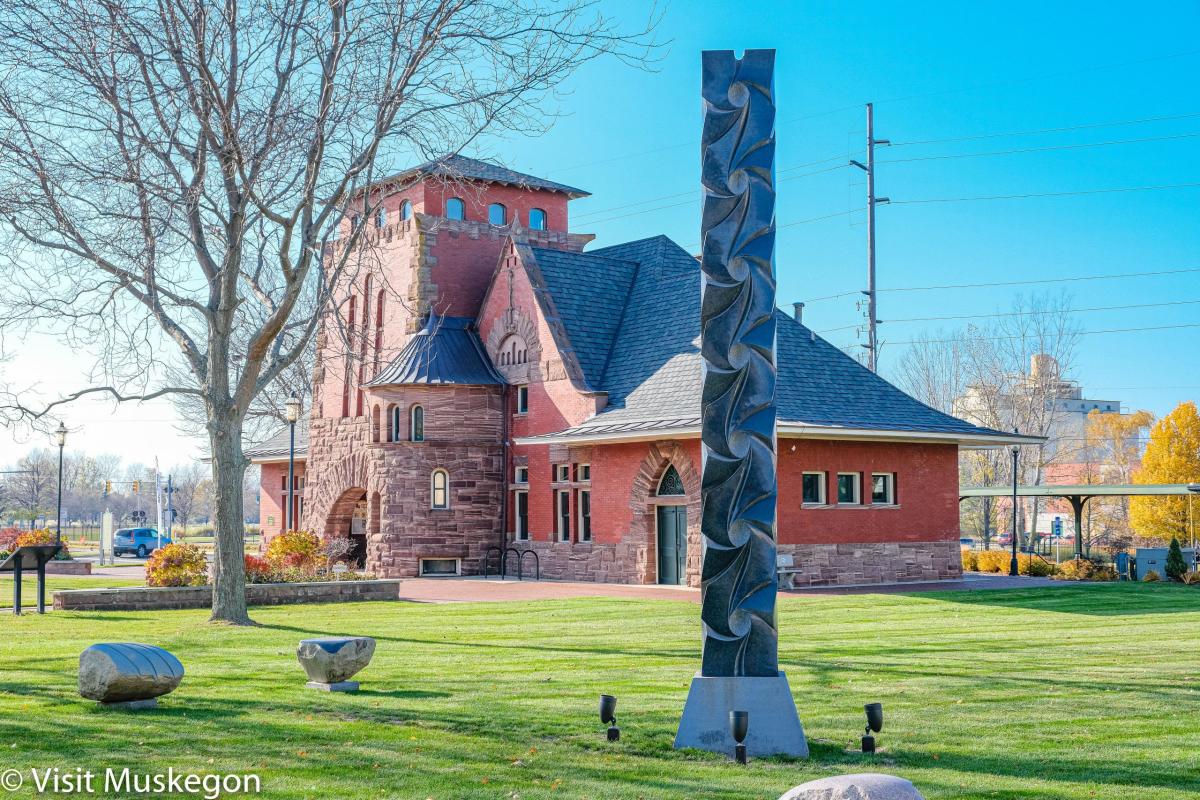 Black granite column sculpture "all my relations" sits on lawn in front of historic brick building.