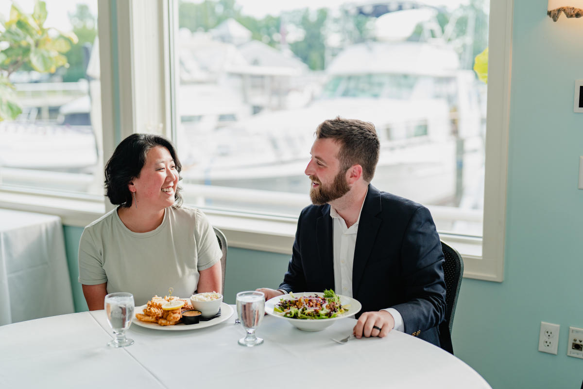 dark haired woman and bearded man smile at one another. They are seated at restaurant with plates of food before them. Docked boats can be seen through the windows behind them