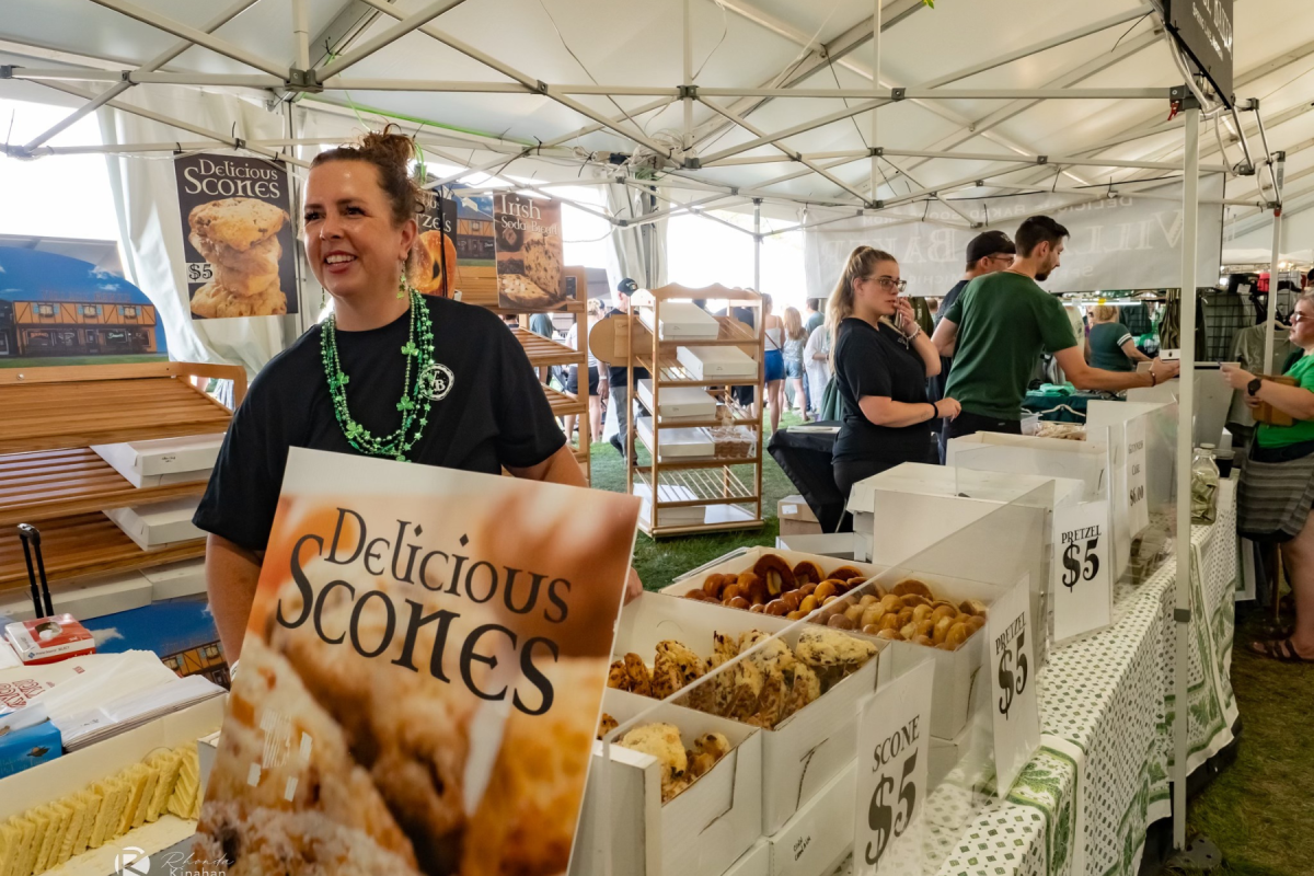 Vendor smiling at a bustling scone booth with signs reading "Delicious Scones" and prices, as customers browse the selections in a tented market area.
