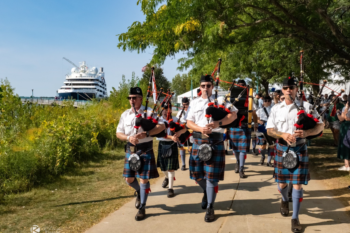 A group of bagpipers in tartan kilts marching in a parade near a large cruise ship docked at Heritage Landing in Muskegon, performing under a clear sky.