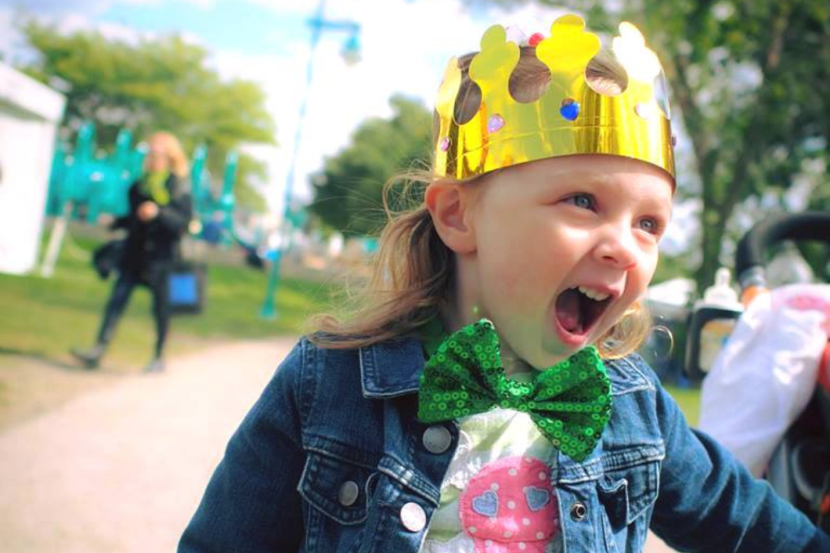 Child wearing a crown and denim jacket, laughing joyously at Michigan Irish Music Festival.