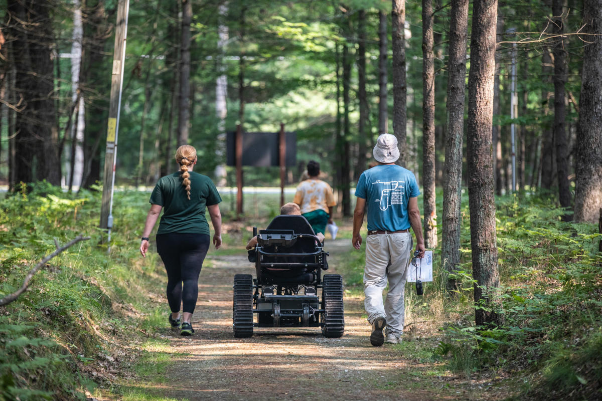 Three people walking on a forest trail, one using a motorized wheelchair.