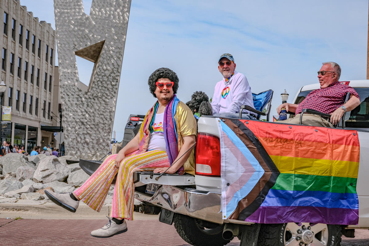 Three individuals on a decorated float featuring a rainbow pride flag, participating in a public event. One person sports dark curly hair and colorful attire, while sitting beside others in casual wear. Modern sculpture visible in the background.