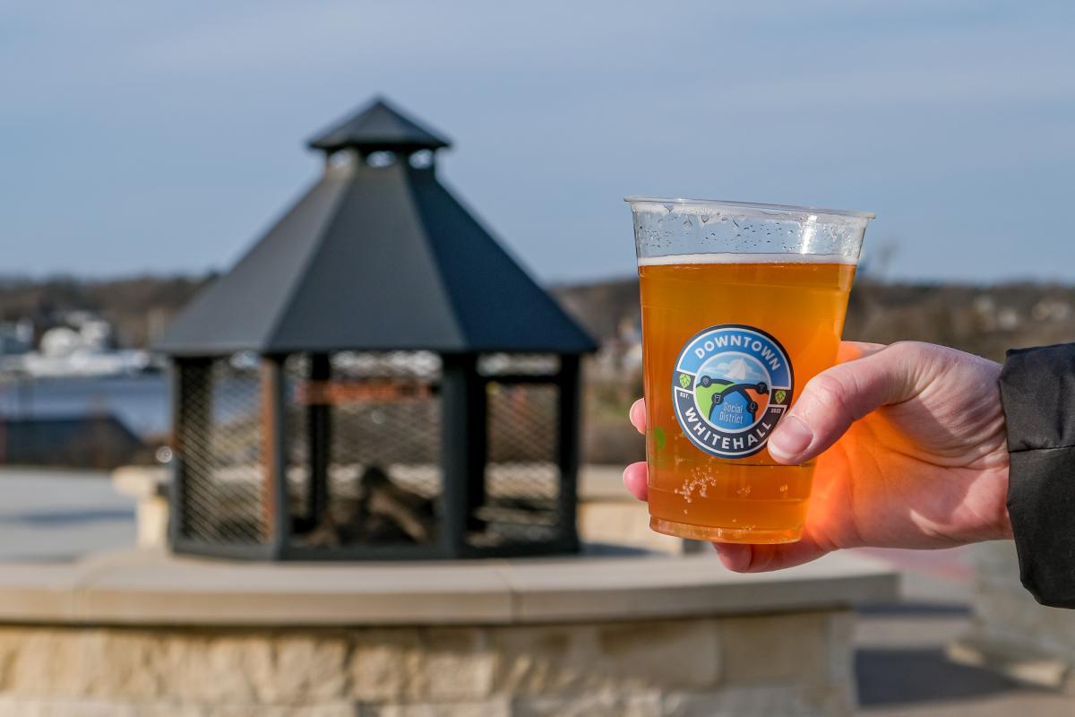 Social District cup of craft beer held up in front of permanent outdoor circular fireplace. Background show a little of white lake, winter blue sky and bare trees.