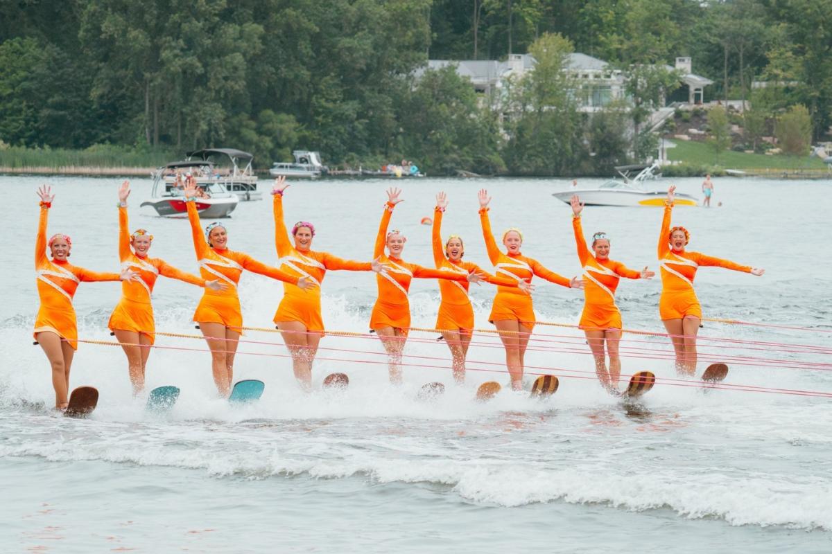 Eight grand city skier performers in orange costumes water skiing in formation on mona lake, with arms raised and a boat visible in the background.