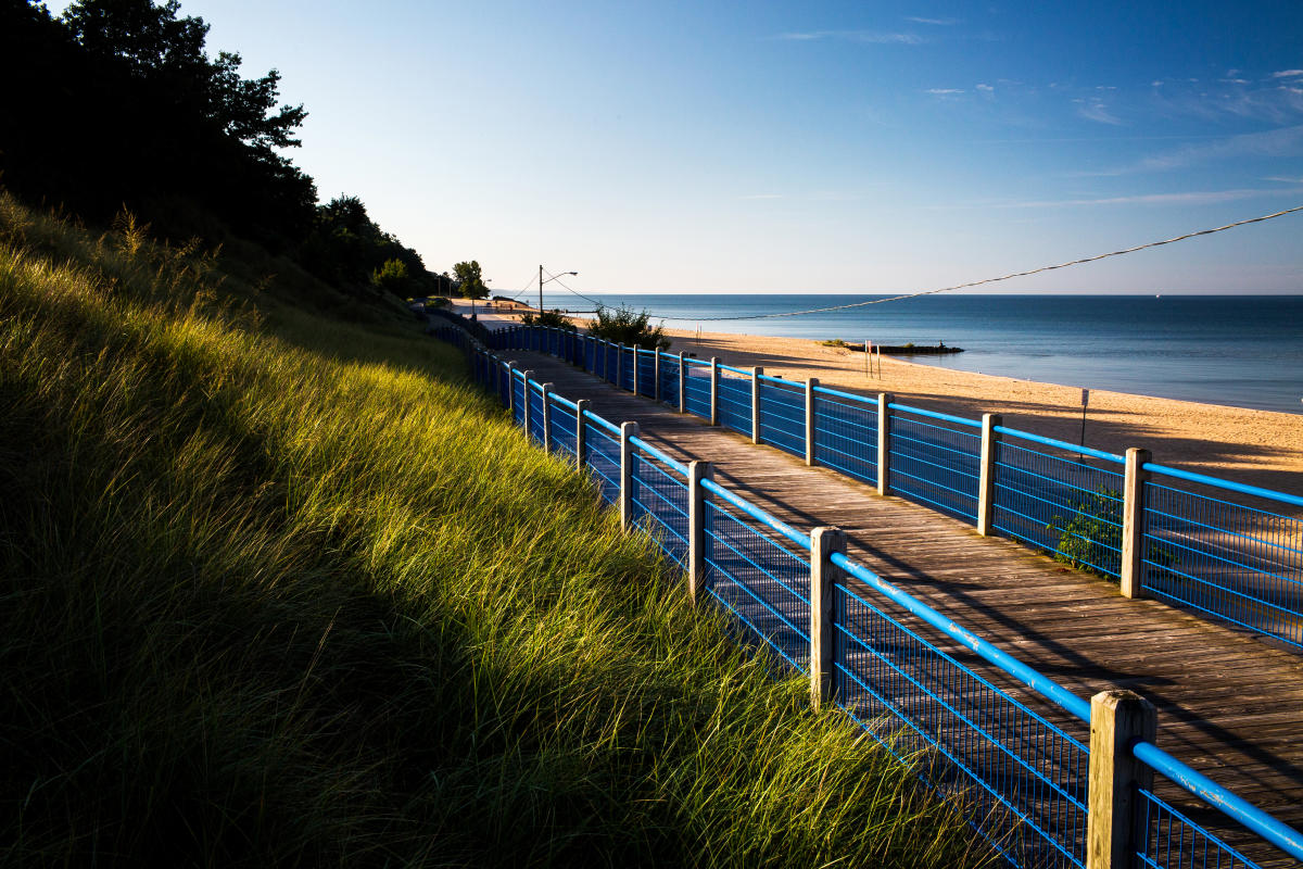 A scenic view of a beach with a long winding pathway lined by a blue railing, overlooking a sandy shore and calm lake Michigan under a clear blue sky.
