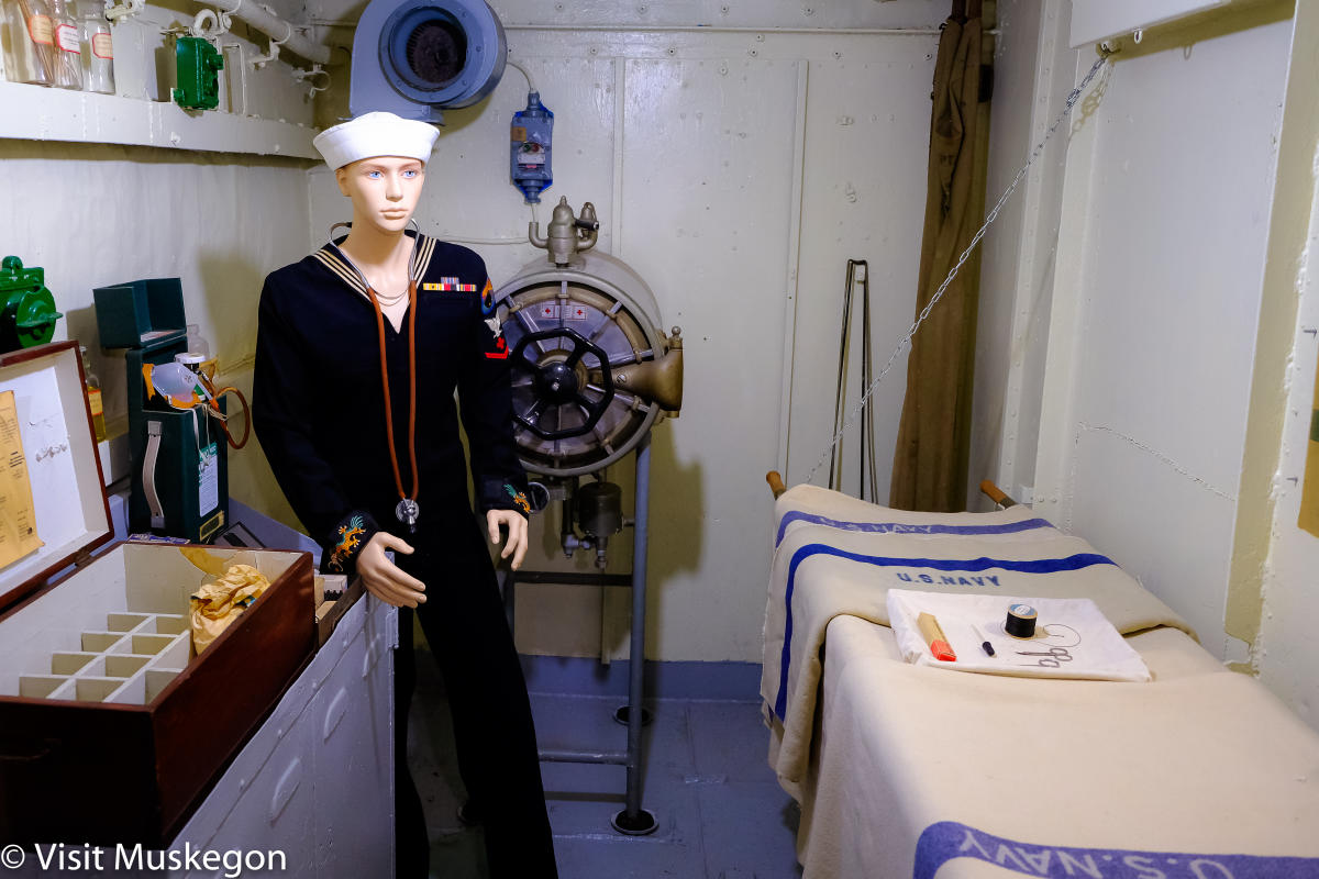 First aid room aboard of USS LST 393 museum shows mannequin in WWII naval uniform standing next to cot in small room with medical supplies