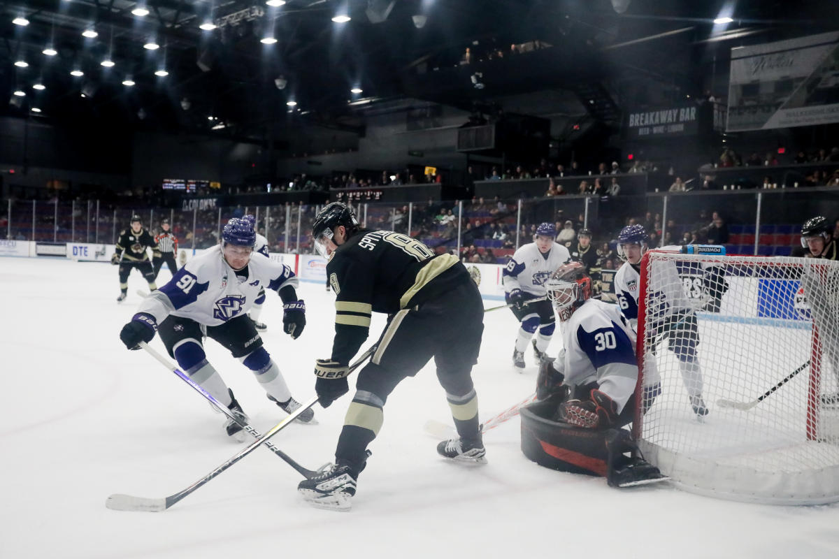 Action shot from a Muskegon Lumberjacks Hockey game at Trinity Health Arena. Two players jostle for puck control near the goal as the goalie prepares to block.
