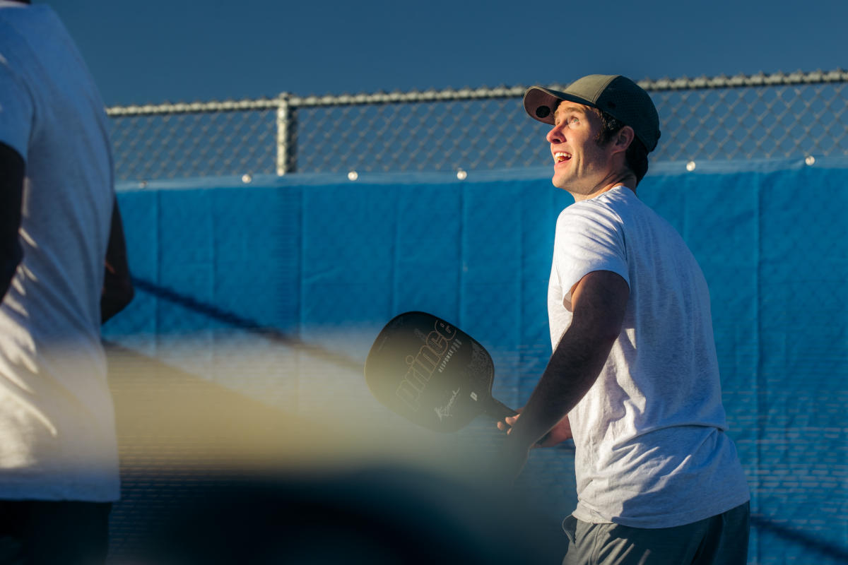 young man in dark baseball cap, white tee shirt and dark shorts smiles as he holds pickleball paddle on outdoor pickleball court