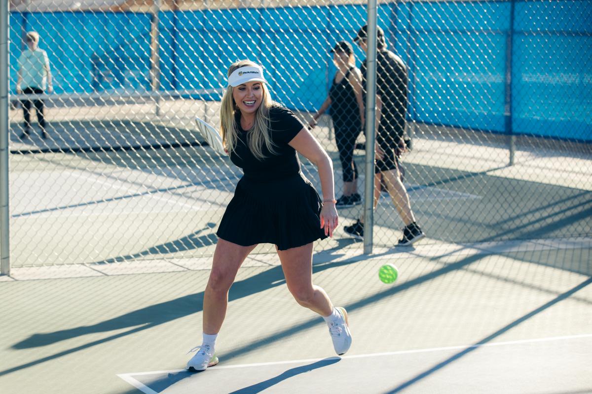 young woman with long blonde hair, wearing white visor cap and black tennis style dress swings at pickleball in outdoor court.