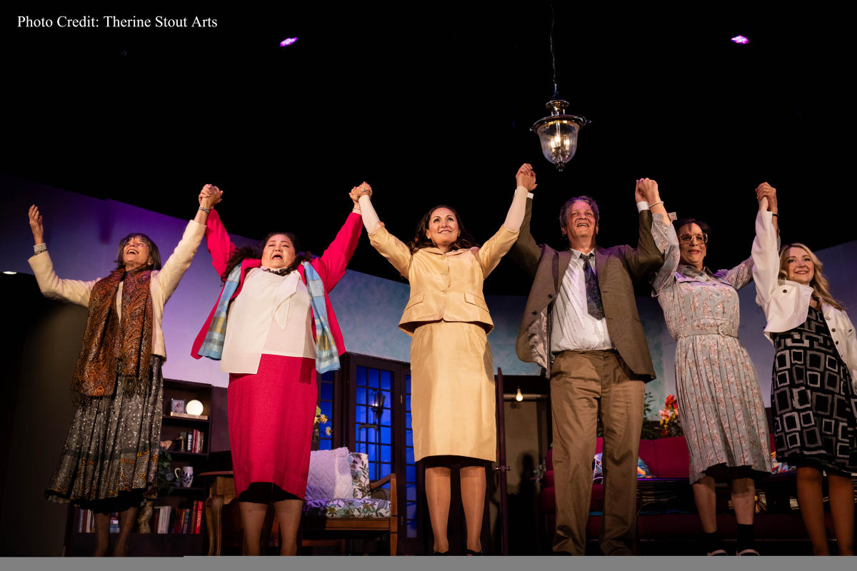 Five actors on Playhouse at White Lake stage taking a bow under a hanging chandelier, with theatrical backdrops visible in the background. They are holding hands raised in the air, smiling broadly in celebration