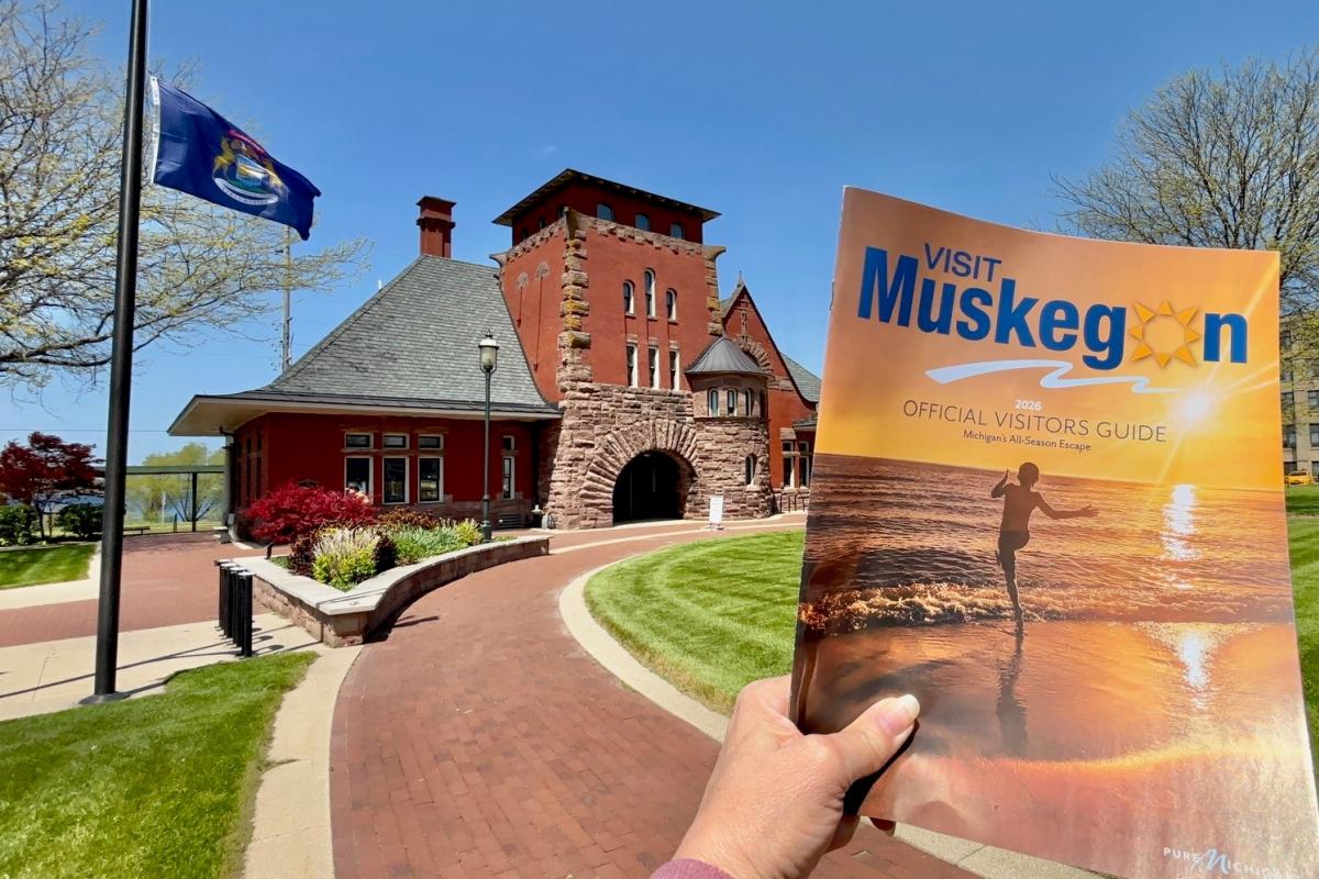 A person holds a "Visit Muskegon" guide with a sunset cover in front of a historic, red-brick building under a clear blue sky, evoking a welcoming feel.