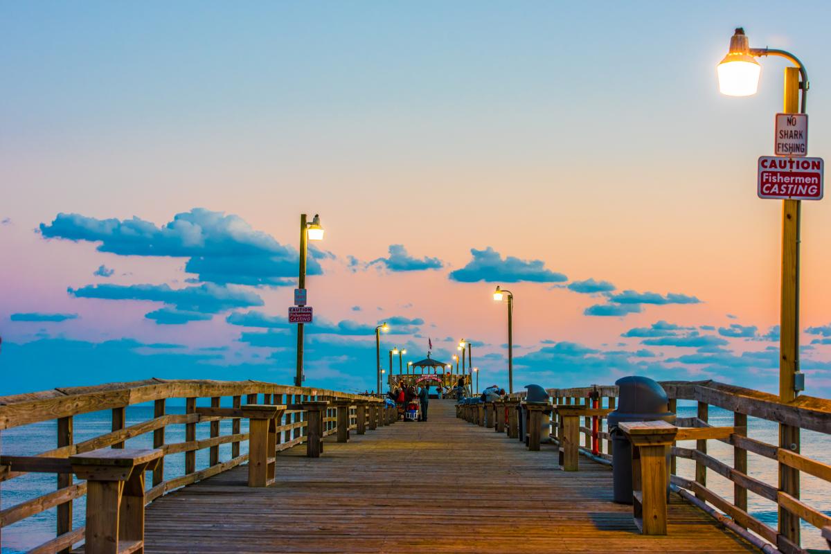 A group of people stand at the end of a long pier in Myrtle Beach illuminated by lamp posts and an orange sky at dusk