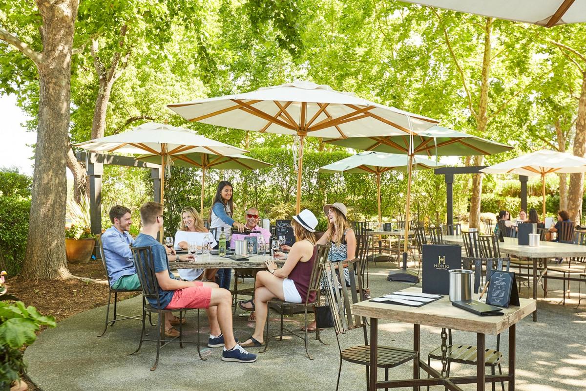 A relaxed outdoor lounge area with umbrellas, seating, and a polished trailer surrounded by Napa Valley vineyards at sunset.