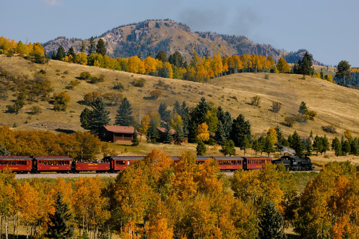 A black steam engine locomotive with red train cars travels down the tracks surrounded by hills, mountains and yellow and green foliage on the trees