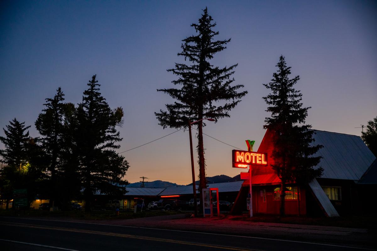 silhouetted trees stand in front of a chalet style building and a sunset with a warm neon sign that reads "motel",