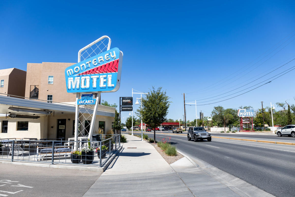 The image shows the exterior of Monterey Motel on a sunny day. The motel features a large retro-style sign with the name brightly displayed. A few cars are driving on the adjacent road, and there are trees and buildings in the background.