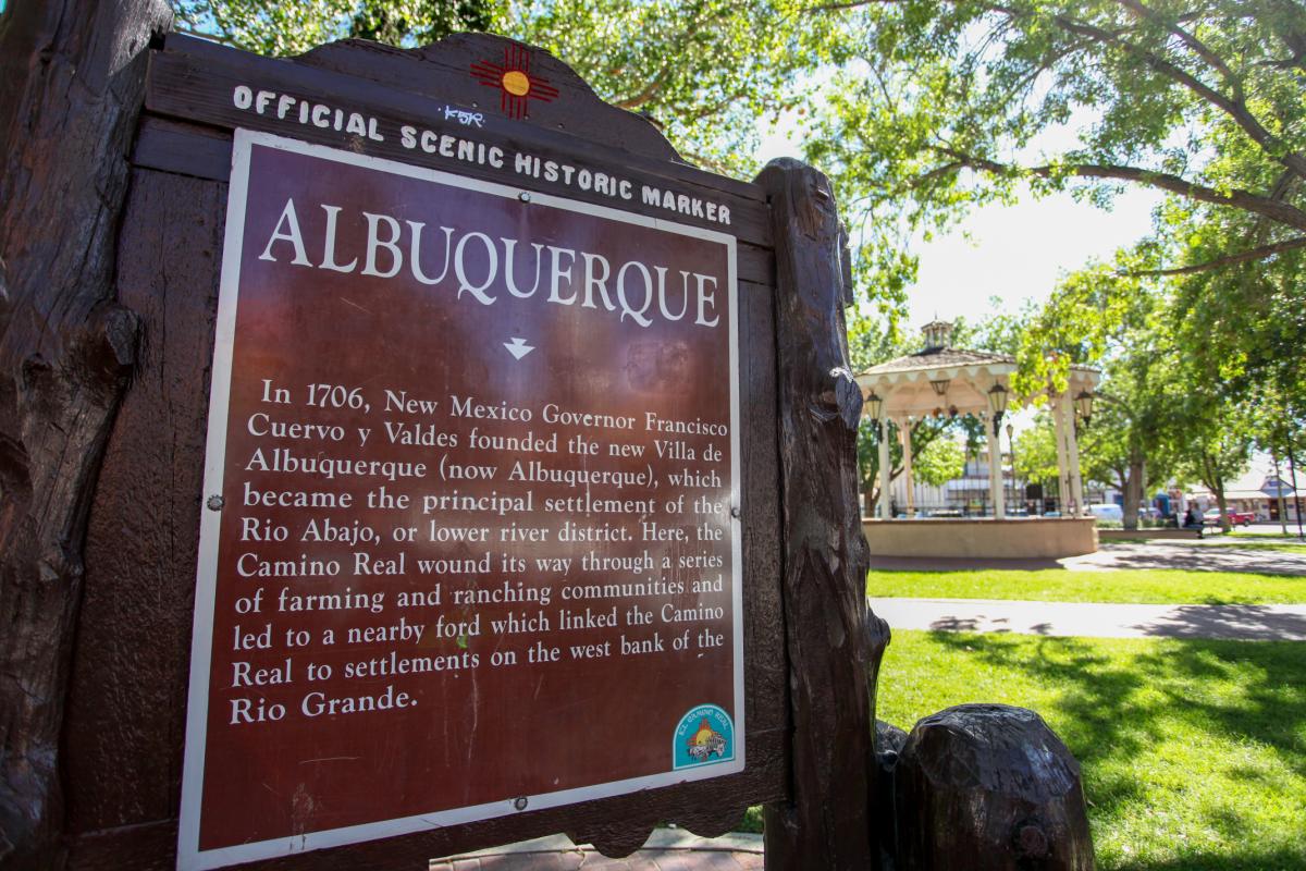 Official scenic historic marker for Albuquerque, detailing its foundation in 1706 by New Mexico Governor Francisco Cuervo y Valdes next to a park with lush trees and a gazebo in the background.