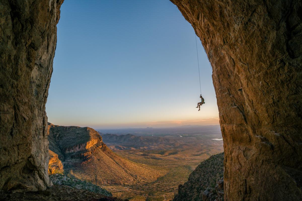 Climber Ben Hanna hangs from a rope anchored into a large arch, overlooking a horizon featuring cliffs and plains