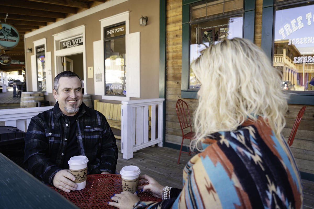 Two people sitting at a table outside a café, holding coffee cups and smiling at each other. The setting includes rustic wooden storefronts with various signs, conveying a quaint, vintage ambiance.