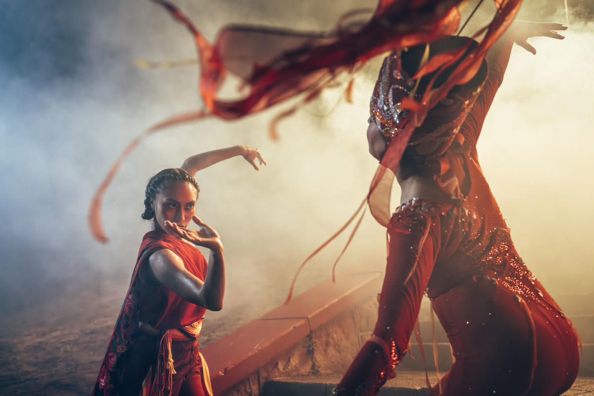 Two individuals in vibrant red attire are dancing gracefully. The scene is dramatic, with flowing fabric and a smoky, atmospheric backdrop.