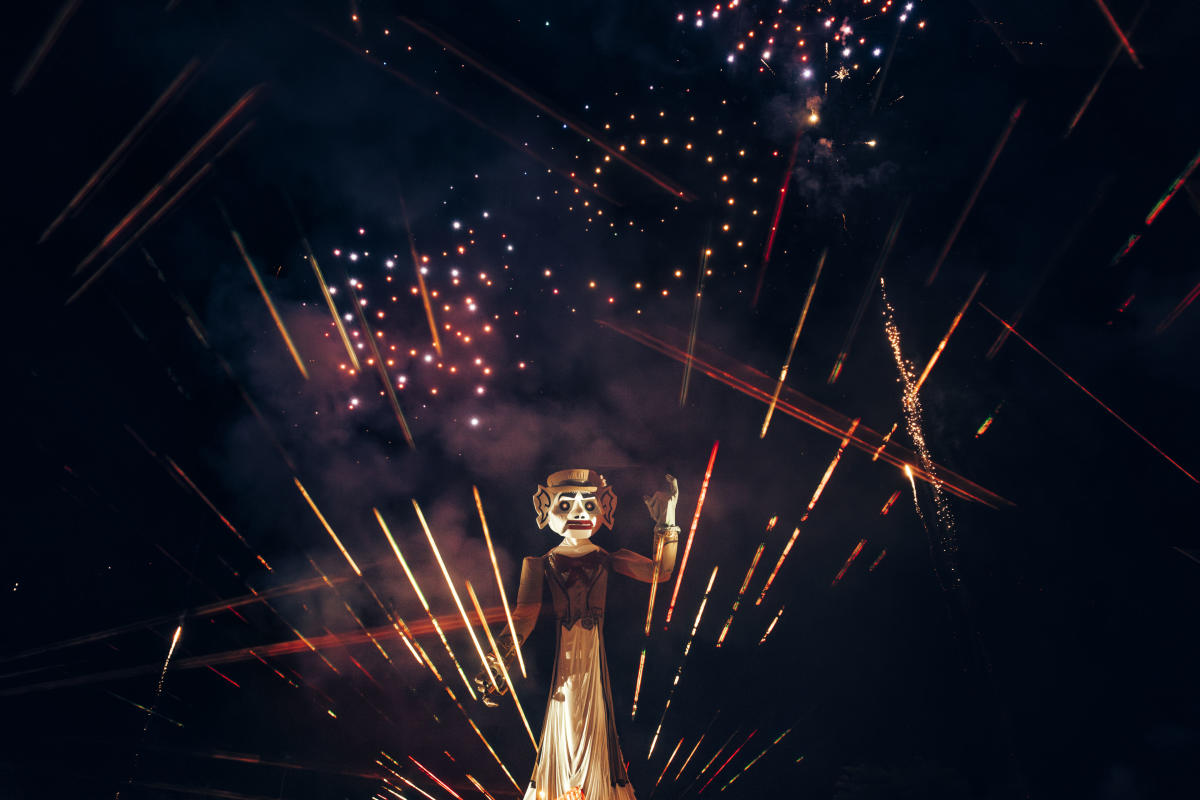 A large effigy of Zozobra is lit up against a night sky filled with colorful fireworks. Bright streaks of light and sparks are scattered around, enhancing the festive atmosphere.