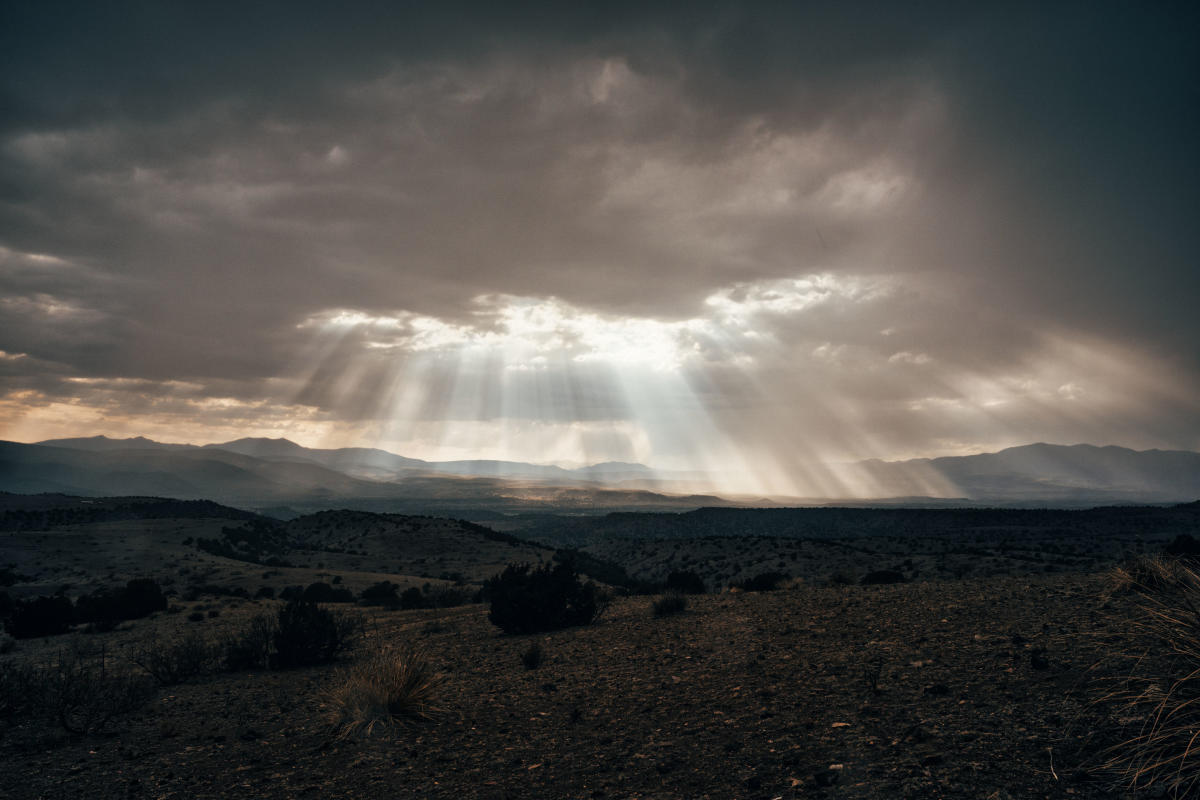 A dramatic landscape featuring a vast desert with sparse vegetation under a cloudy sky. Sunbeams pierce through the clouds, casting light over the terrain. Mountains are visible in the distant background.