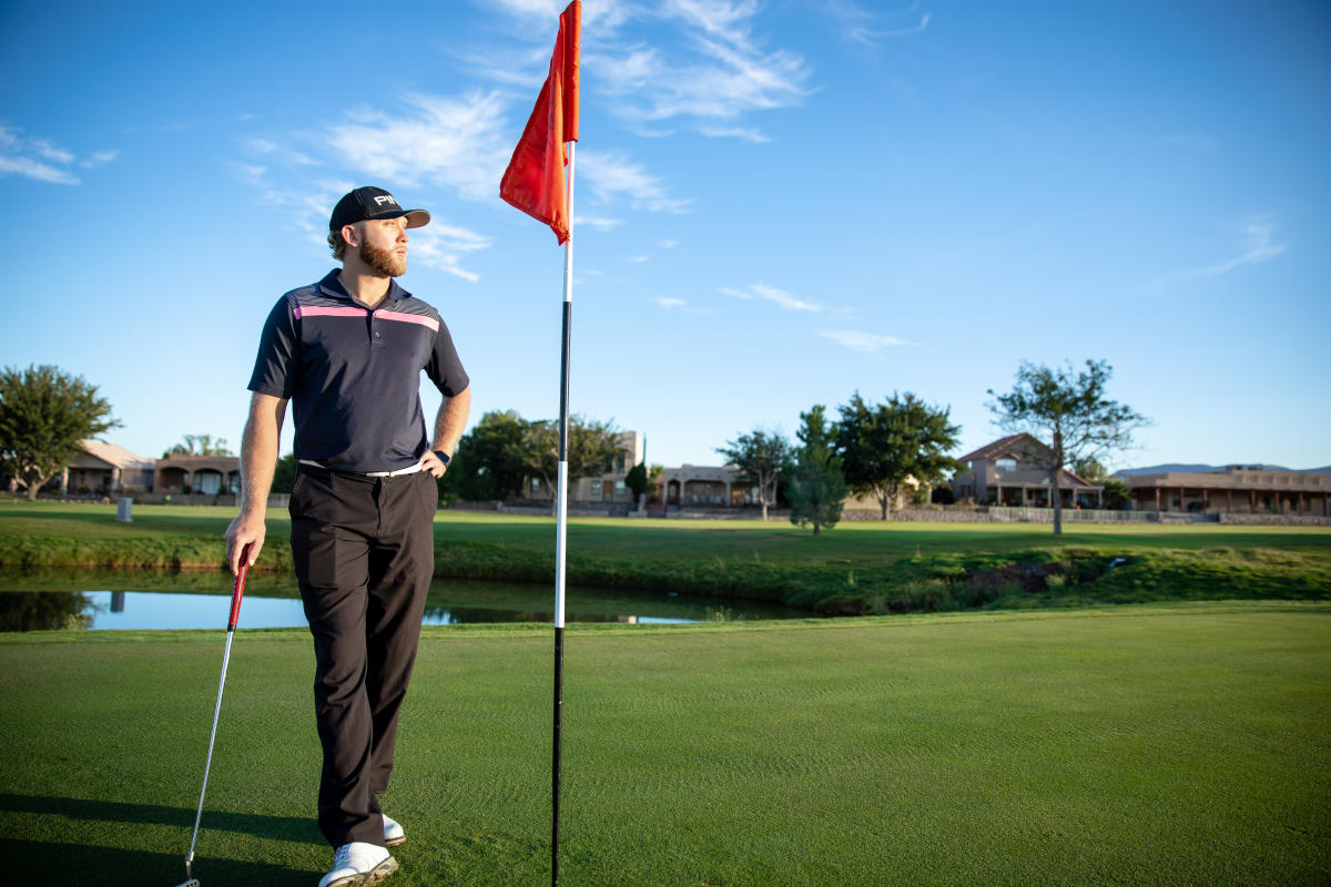 A person stands next to a golf flag on a green at a golf course, holding a putter, with residential buildings and a pond in the background.