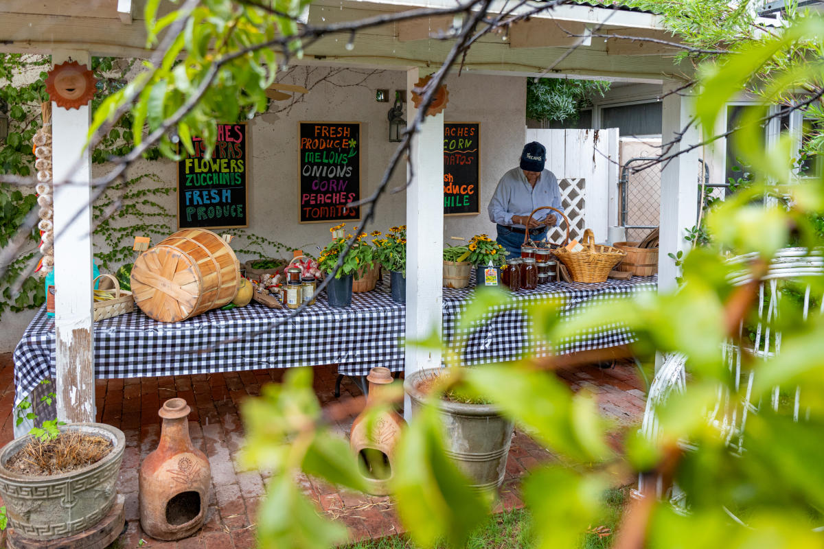 A person stands at a produce stand under a sheltered area, surrounded by baskets and jars of pickled goods. Signs list items such as fresh produce, melons, and tomatoes.