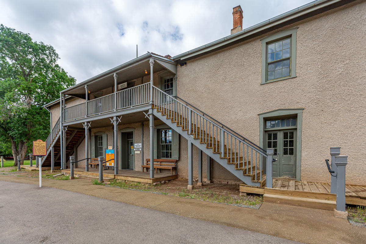 Historic two-story adobe building with an external staircase and wraparound balcony. The sky is partly cloudy, and trees are visible in the background.