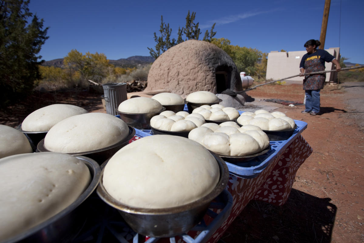 Trays full of bread dough sit on a table outside, as a person in the background inserts bread into a traditional horno with a long utensil