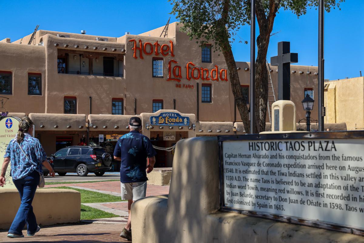A sunlit street scene featuring the adobe-style Hotel La Fonda in Taos. The sign for Hotel La Fonda is prominently displayed on the building. Two individuals walk along a path lined with trees. In the foreground, there is a plaque with text about the historic Taos Plaza.