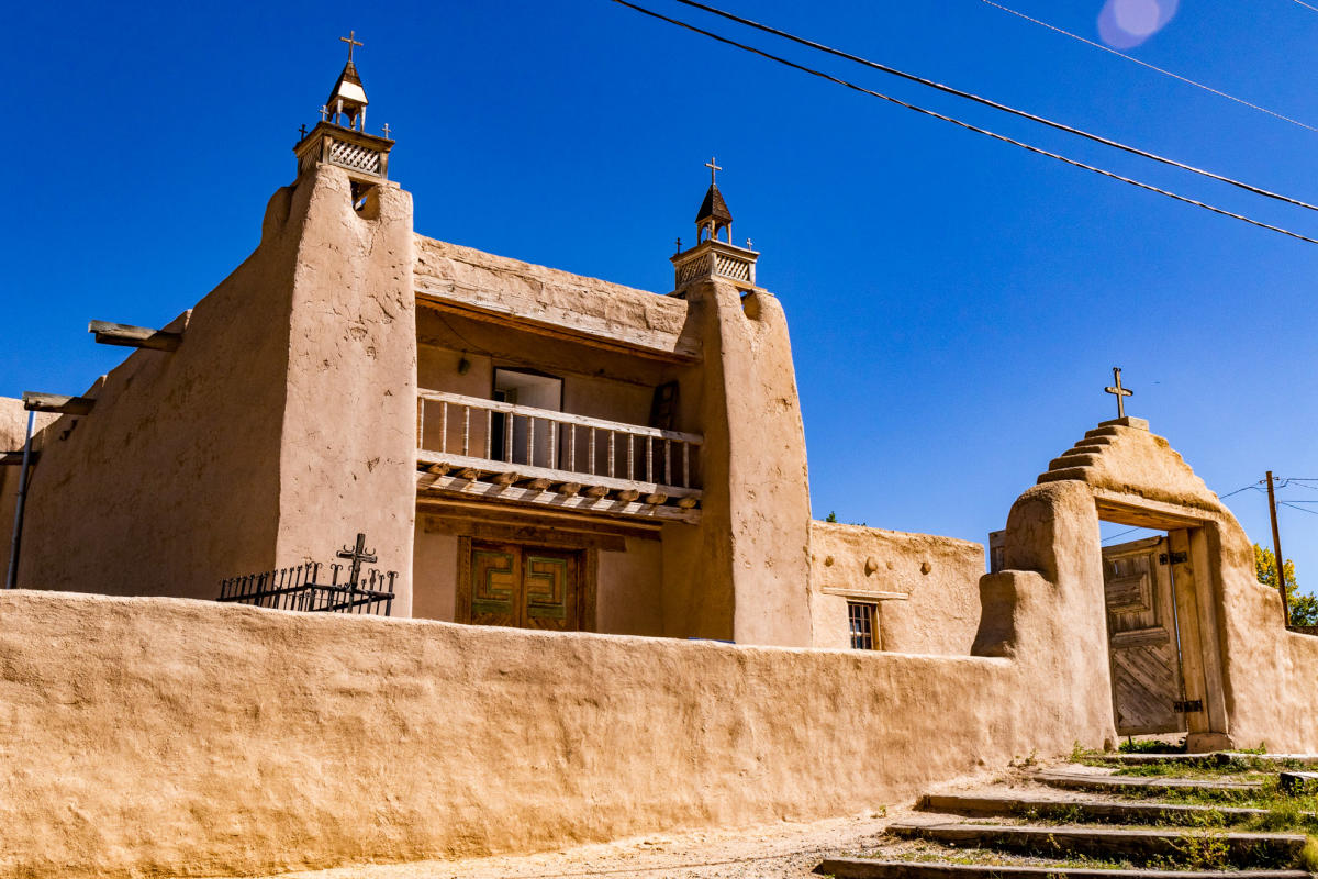 A historic adobe church with thick walls and two bell towers under a clear sky, featuring wooden doors and a small entrance arch with a cross on top.